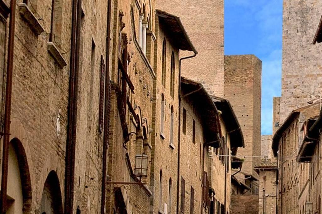 A narrow street with historic stone buildings, arched doorways, and a prominent tower in the background.