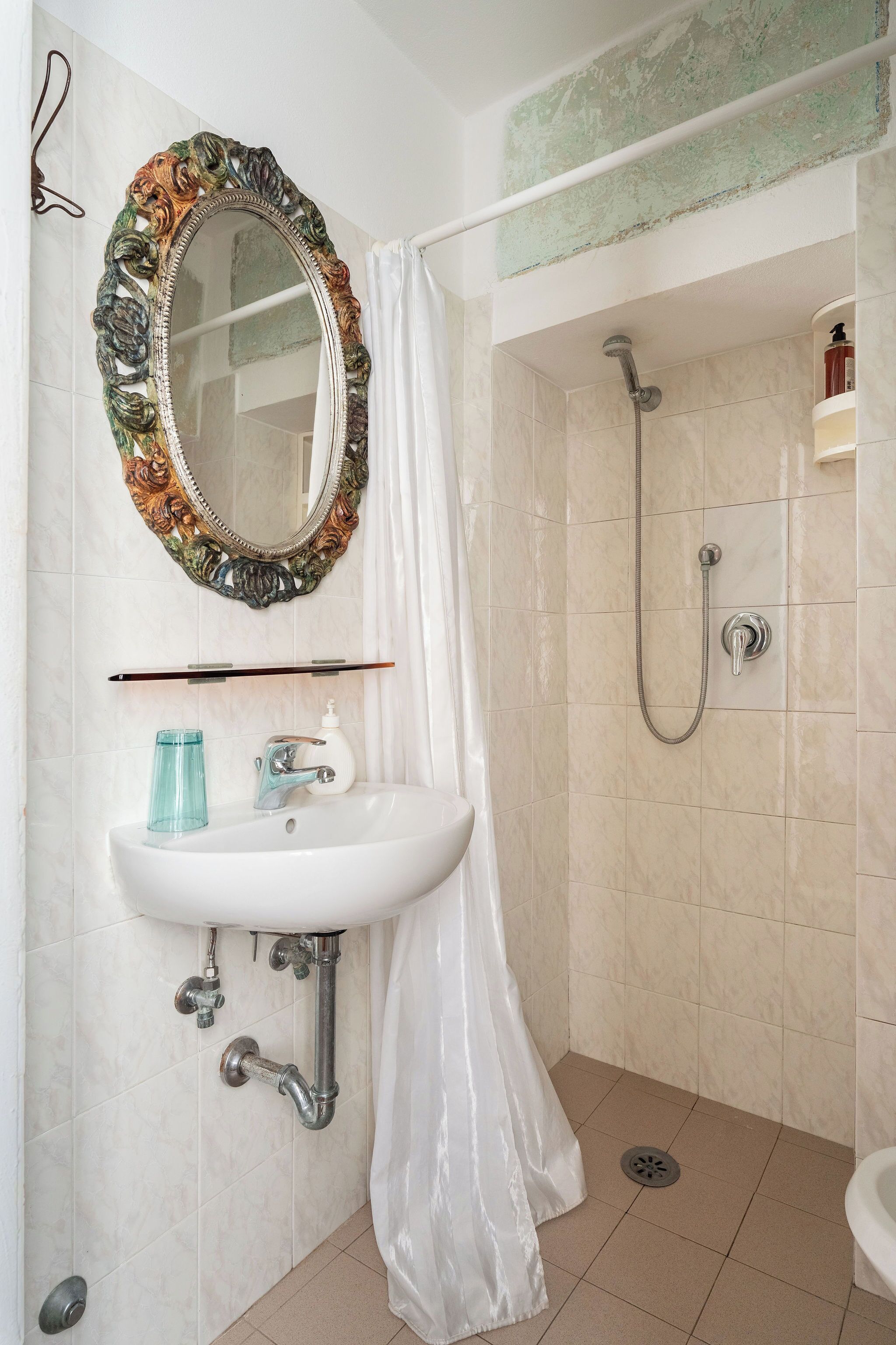 Une salle de bain avec un lavabo blanc, un miroir doté d’un cadre orné, une douche avec un pommeau de douche à main et un rideau de douche blanc.