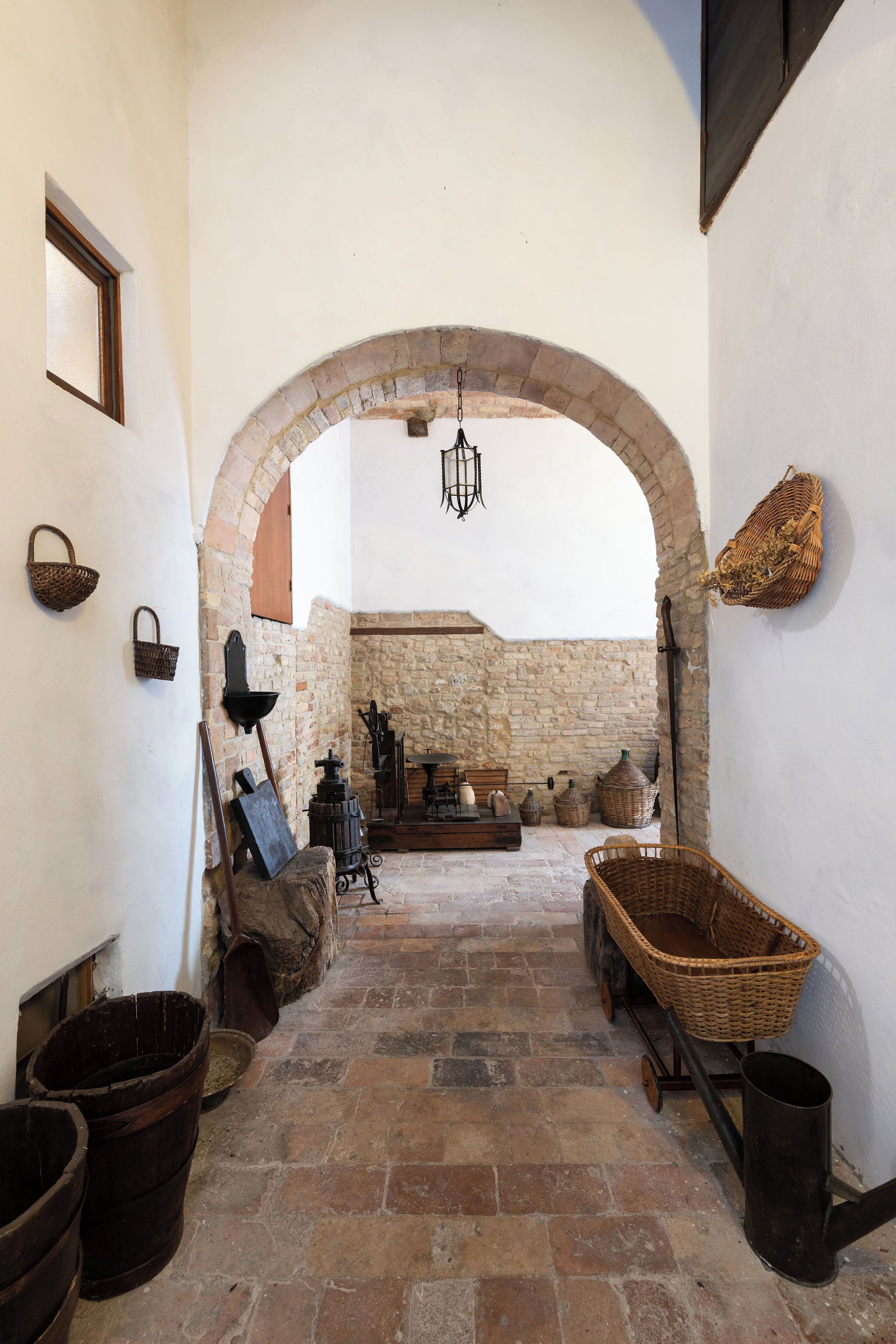 An arched corridor with stone walls, a hanging light fixture, and rustic furniture including a wooden cart and baskets.