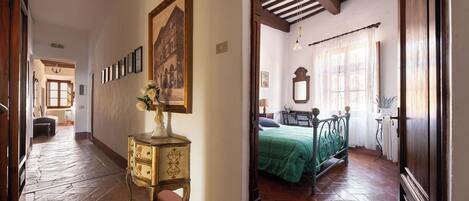 Un pasillo con piso de madera, un aparador antiguo, un cuadro enmarcado y una cama con colcha verde en la habitación contigua.