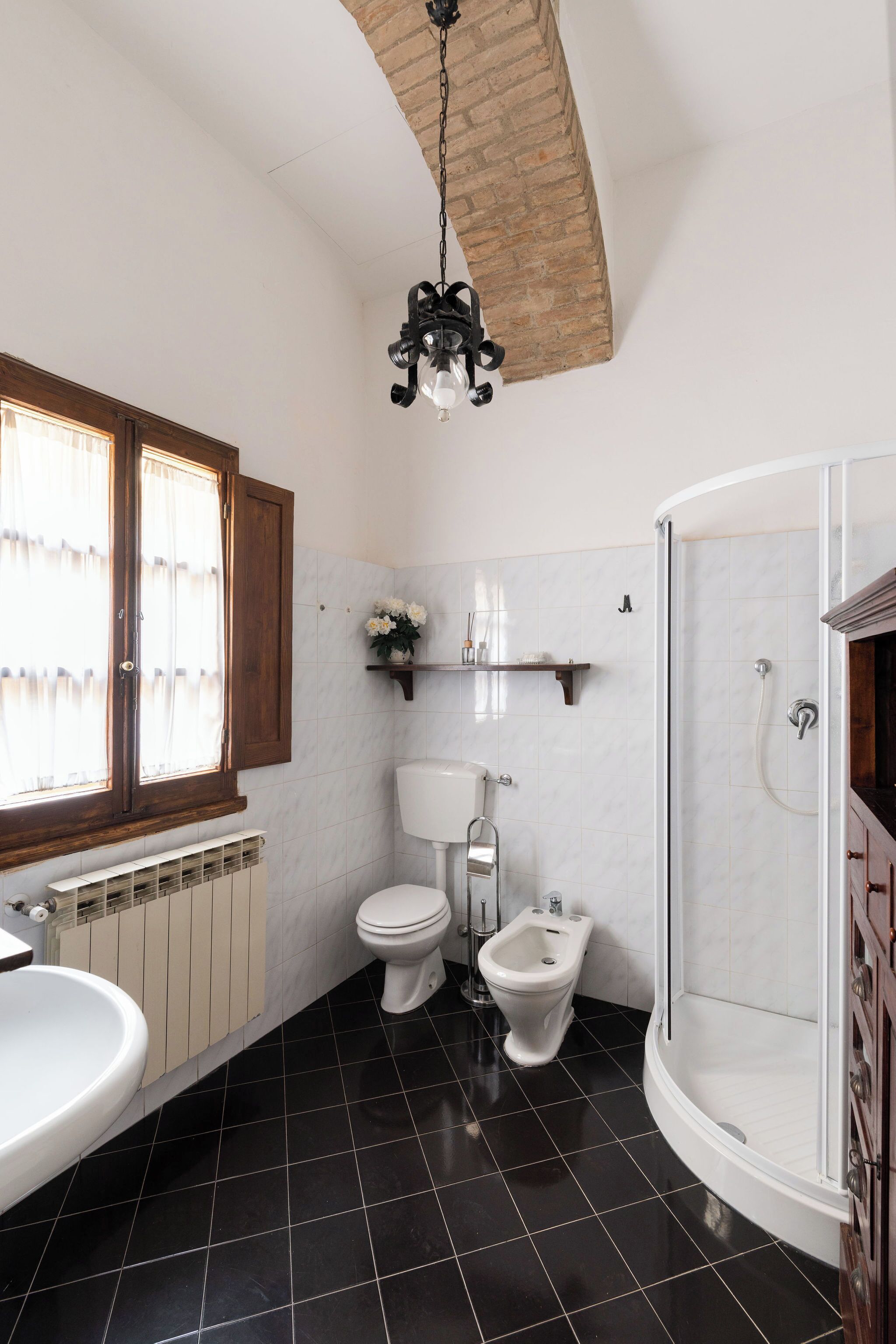Un bagno con un water bianco, un bidet e una doccia. Si vede una finestra con tende, un termosifone e uno scaffale con articoli da toilette.
