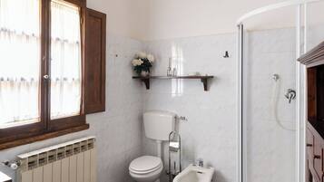 Un bagno con un water bianco, un bidet e una doccia. Si vede una finestra con tende, un termosifone e uno scaffale con articoli da toilette.