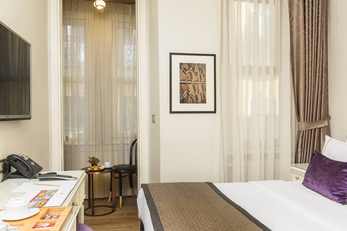A hotel room with a bed, a desk with a telephone, a chair, and a small table with a cup and a plate of fruit.