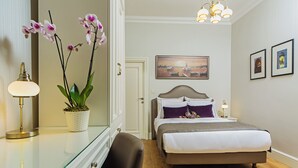 A hotel room with a bed, a desk with a lamp, a potted orchid, and a painting on the wall.