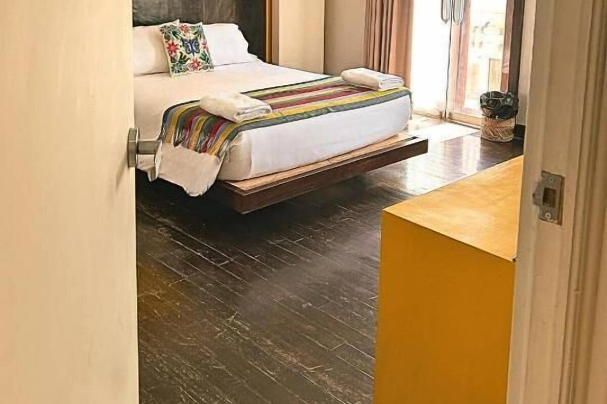 A hotel room with a bed, a wooden floor, a yellow accent wall, and a sliding glass door leading to a balcony.