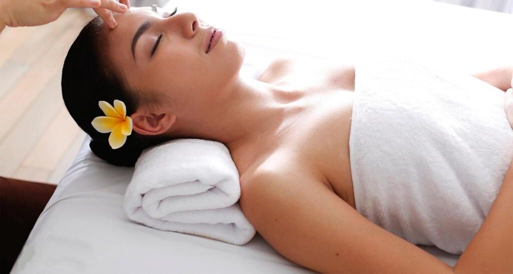 A person lying on a bed with a white towel and a flower in their hair.