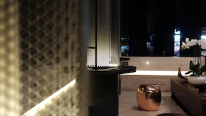 Et moderne interiør med en struktureret væg, en cylindrisk søjle med perforeret design, og en træbænk med en potteplante.