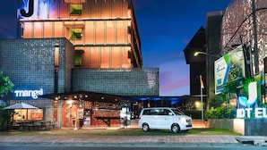 Et moderne hotel med et tydeligt 'J'-skilt, en café ved navn 'Mangost' og en hvid varevogn parkeret foran.