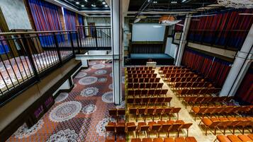Ein Theater mit Reihen orangefarbener Sessel, einem gemusterten Boden und roten Vorhängen.