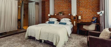A hotel room with a brick wall, a large bed with white bedding, a wooden nightstand, a chair, and a desk with a lamp.