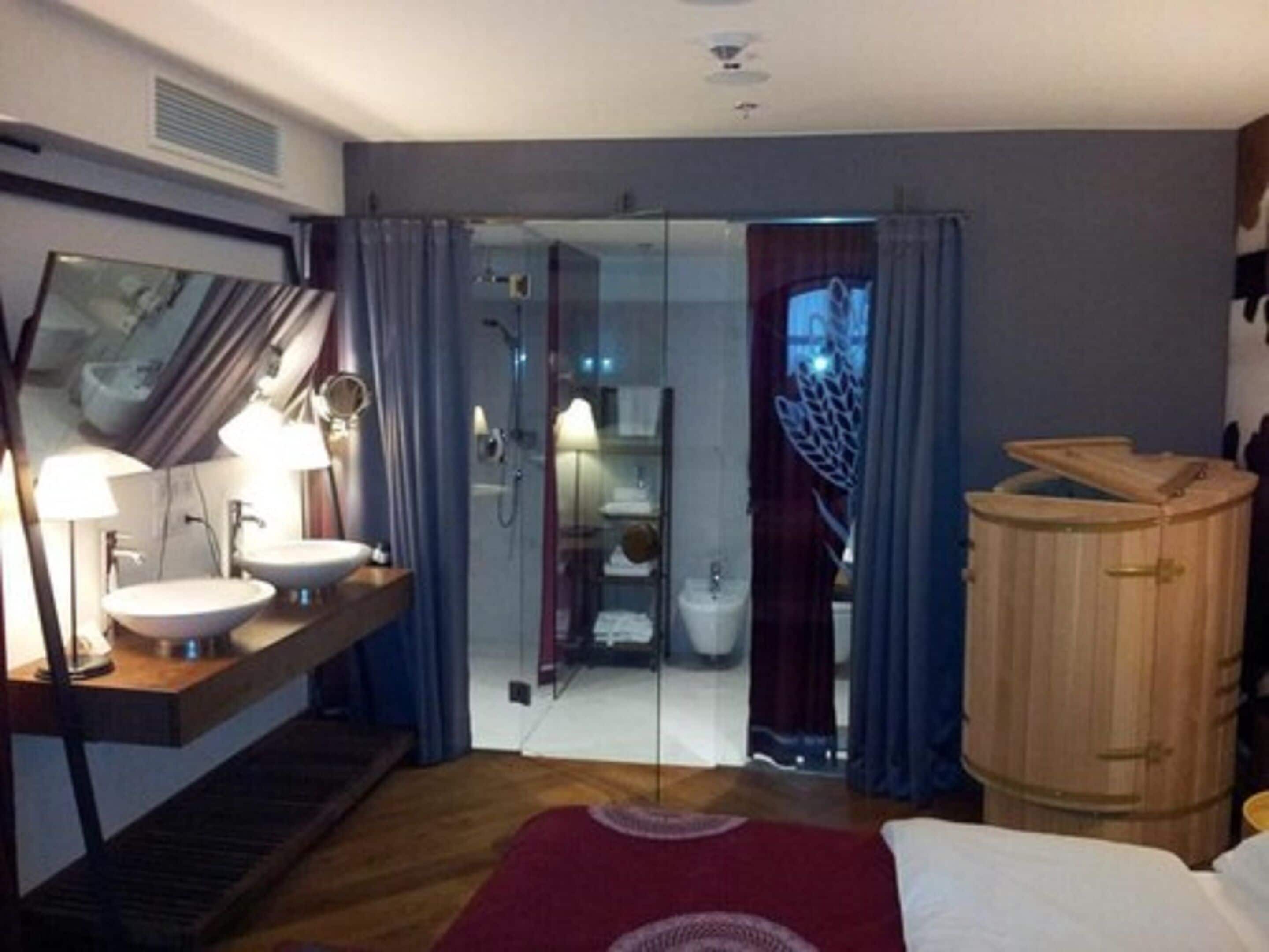 A hotel room with a wooden cabinet, a large mirror, a bed with a red rug, and a bathroom visible through a glass door.