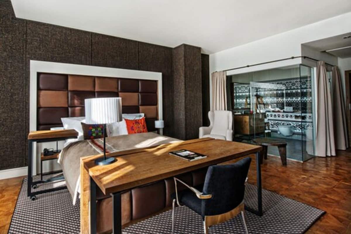 A modern hotel room with a large bed, a wooden desk, a chair, and a glass partition with a view of the city.
