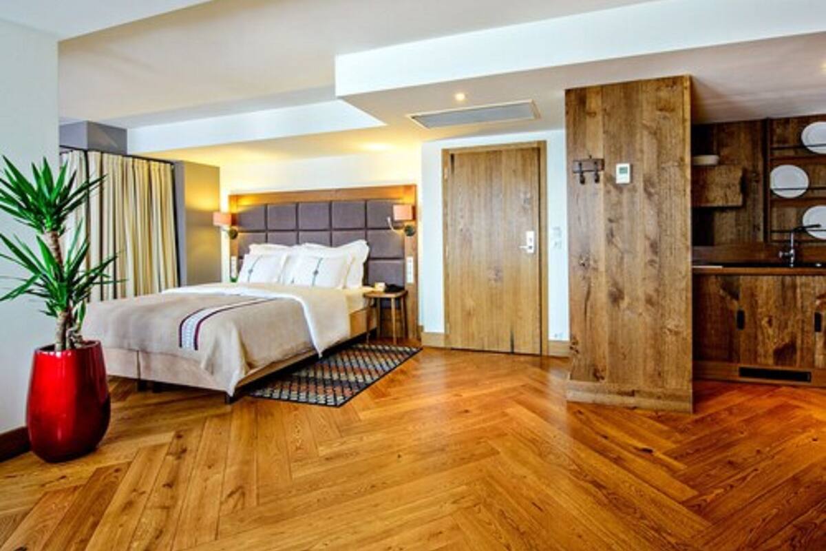 A modern hotel room with a large bed, wooden flooring, a potted plant, and a wooden kitchen area.