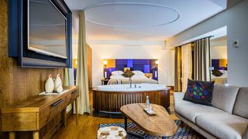 A hotel room with a wooden desk, a round coffee table, a sofa, a bed with a headboard, and a TV mounted on the wall.