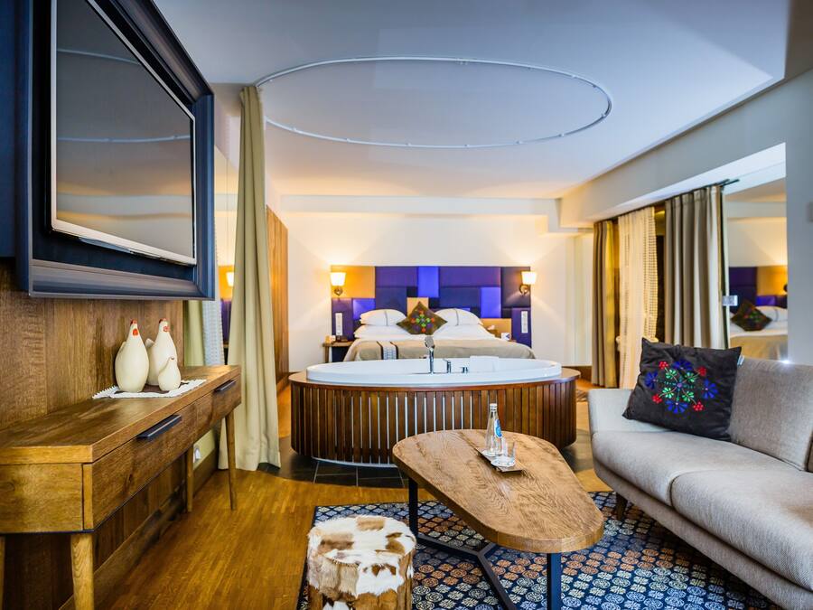 A hotel room with a wooden desk, a round coffee table, a sofa, a bed with a headboard, and a TV mounted on the wall.