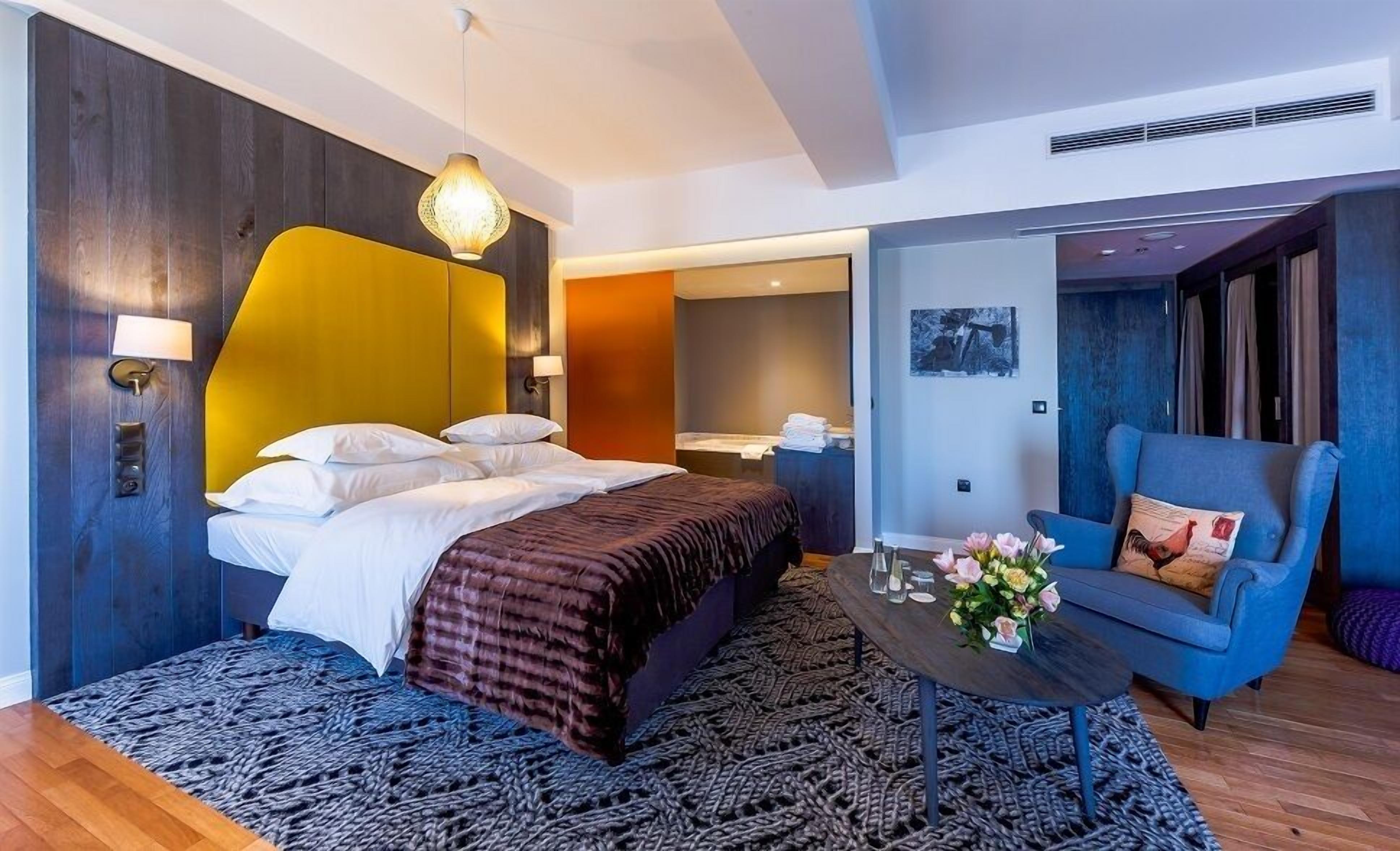 Ein modernes Hotelzimmer mit einem großen Bett, einem kleinen Tisch mit Blumen, einem blauen Sessel und einer gelben Akzentwand.