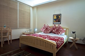 A neatly arranged bedroom with a wooden bed, a desk, a chair, a bedside table with a lamp, and a window with a view. - Colonel's Retreat At The Airport (New Delhi)