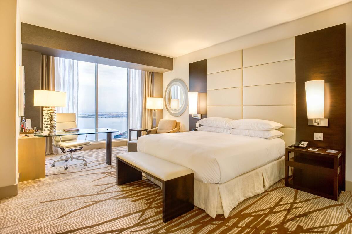 A hotel room with a large bed, a desk with a chair, a bench, a round mirror, and a nightstand with a lamp.
