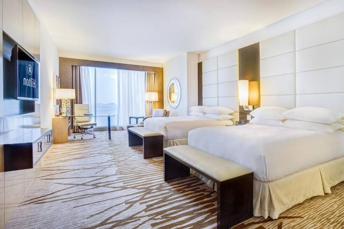 A hotel room with two beds, a desk, a TV, and a view of the outdoors.