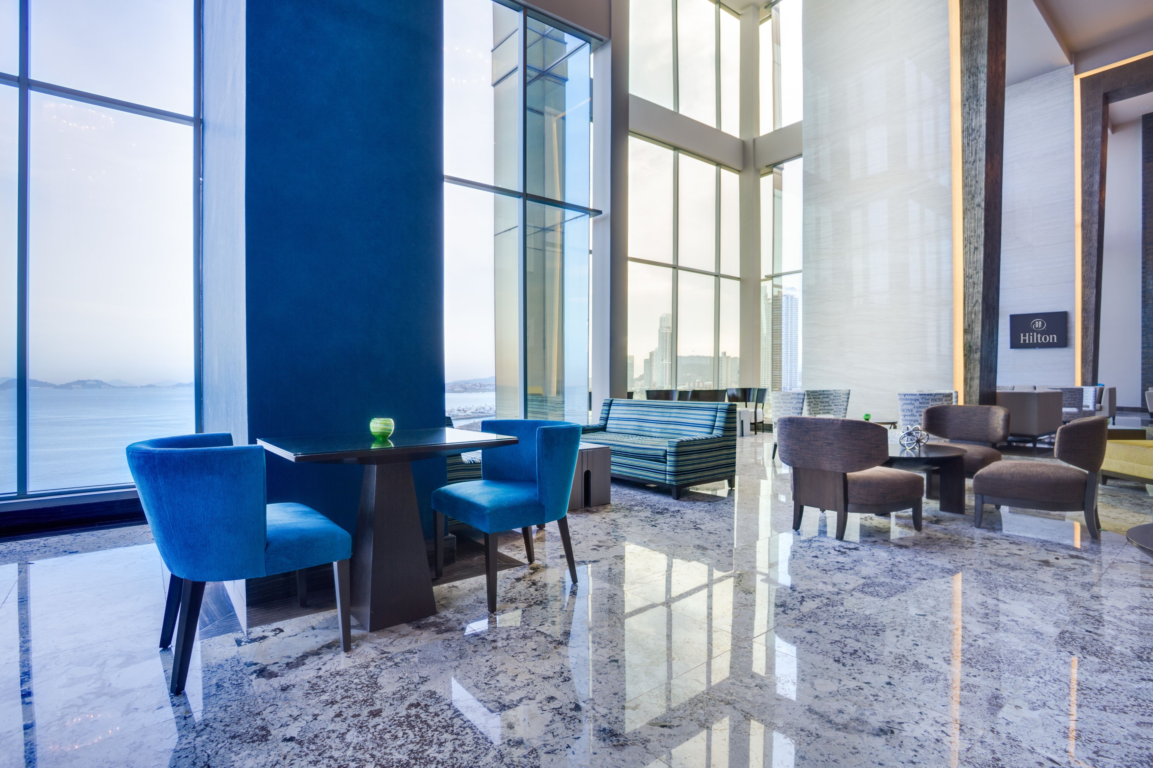 Un hall d’hôtel moderne doté d’une grande fenêtre offrant une vue sur le paysage urbain. On y trouve des chaises bleues, une petite table et un espace salon avec des chaises marron.