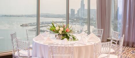 A round table with white chairs and a floral centerpiece, set against a large window overlooking a cityscape and water.