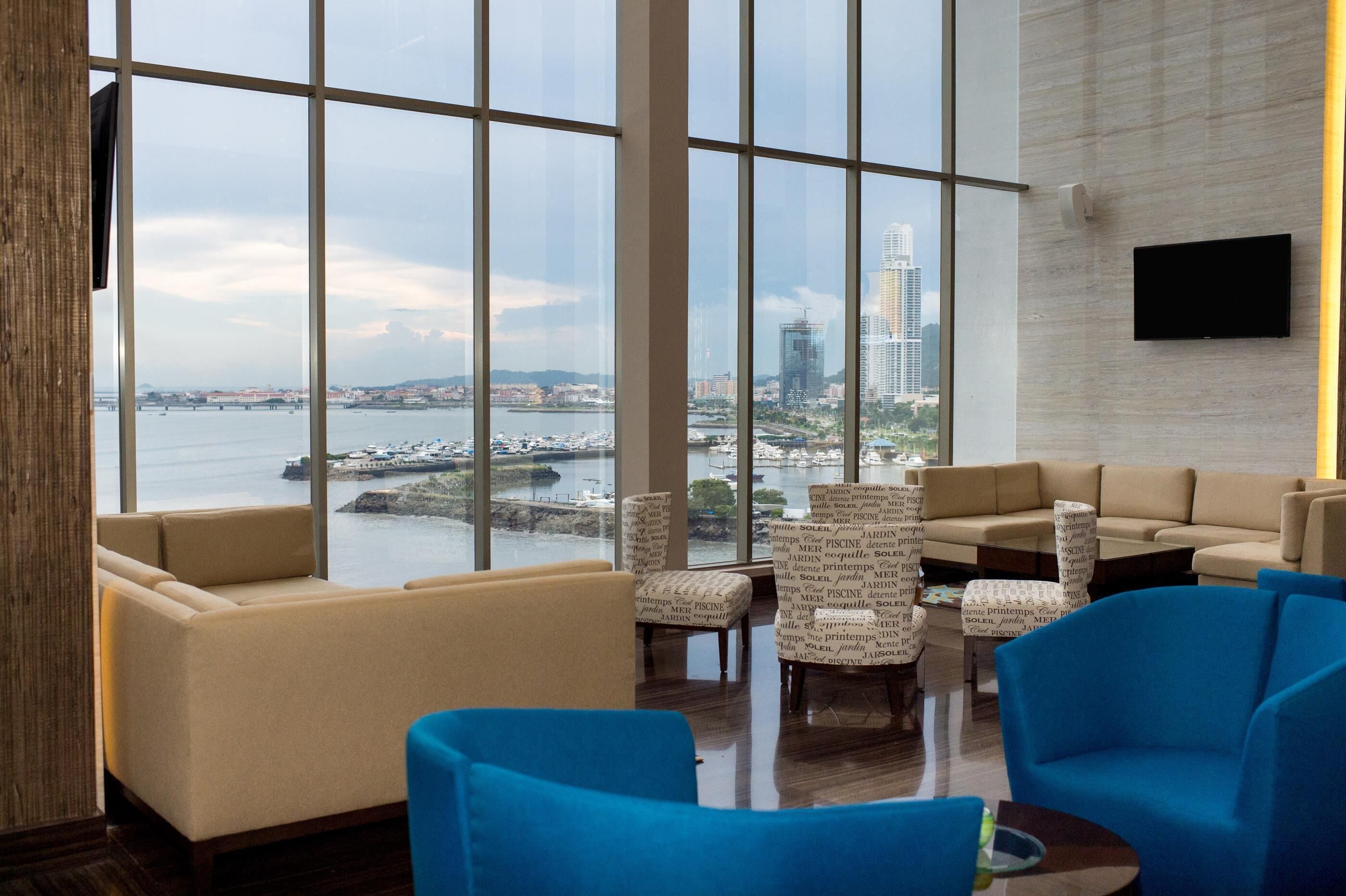 A modern hotel lobby with large windows, comfortable seating, and a flat-screen TV on the wall.