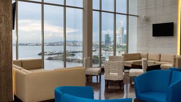 A modern hotel lobby with large windows, comfortable seating, and a flat-screen TV on the wall.