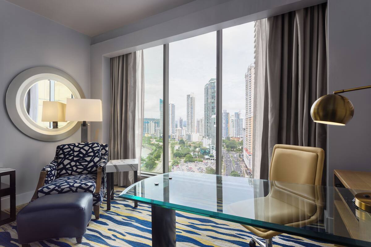A modern hotel room with a glass table, two chairs, a round mirror, and a view of the cityscape.