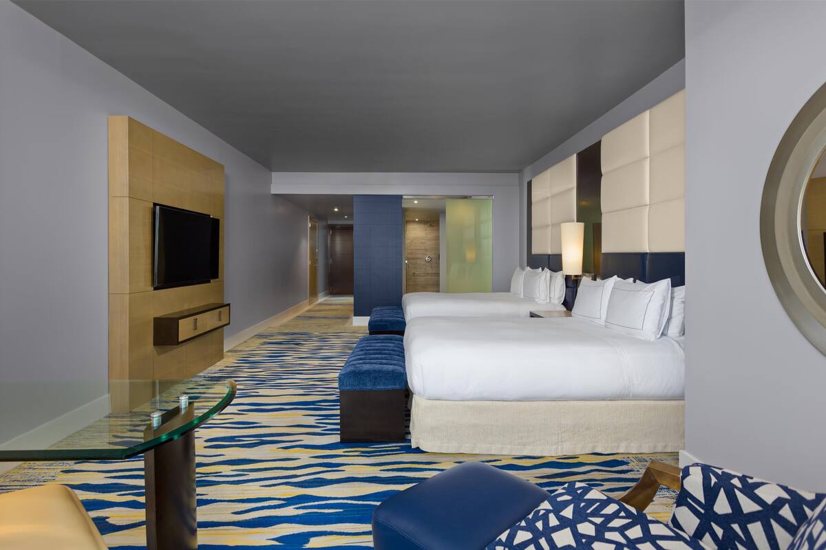 A modern hotel room with a large bed, a TV, a glass table, and a patterned carpet.