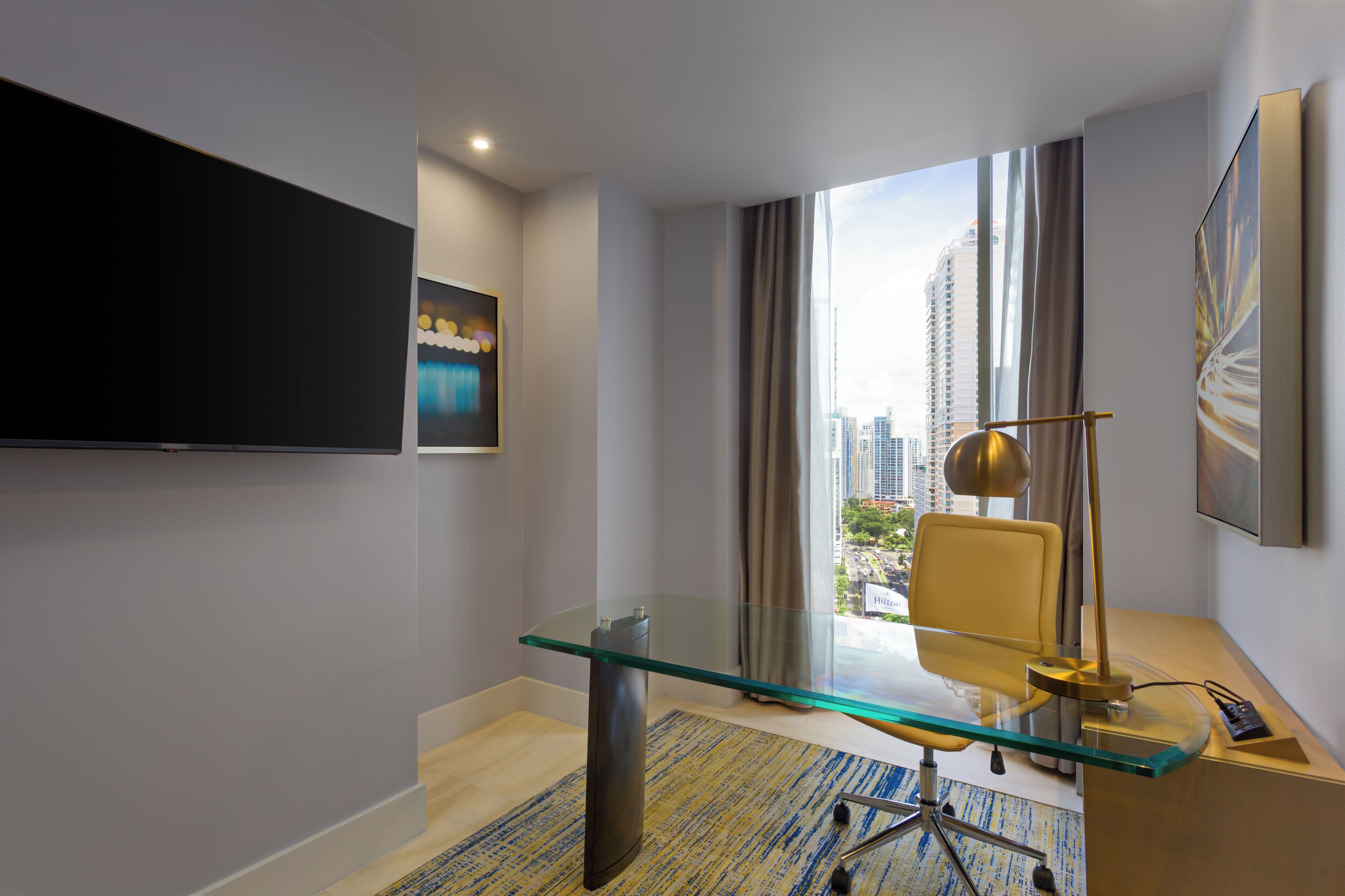 Une chambre d’hôtel moderne dotée d’un bureau en verre, d’une chaise, d’un téléviseur à écran plat, d’un tableau et offrant une vue sur la ville.