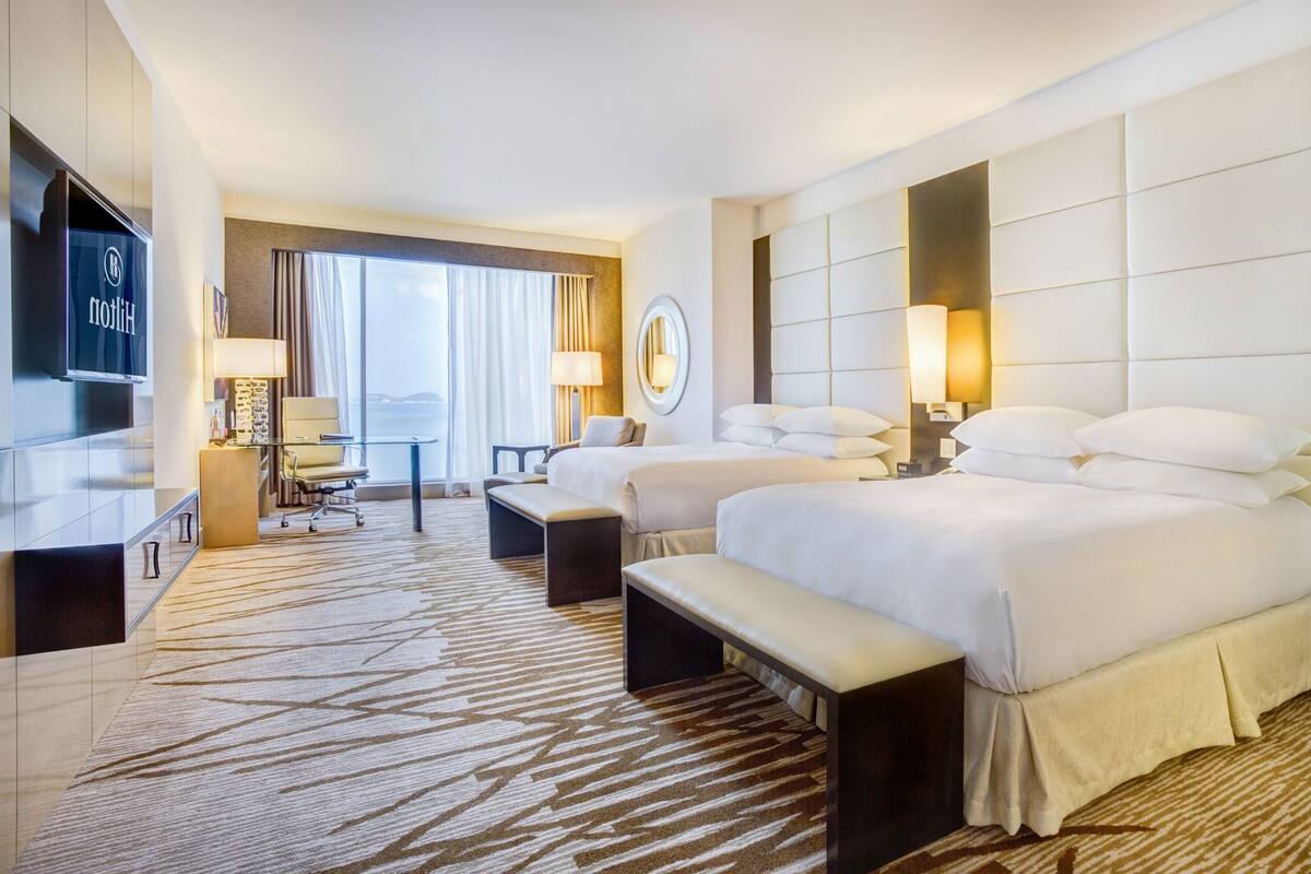 A hotel room with two beds, a desk, a TV, and a view of the outdoors.