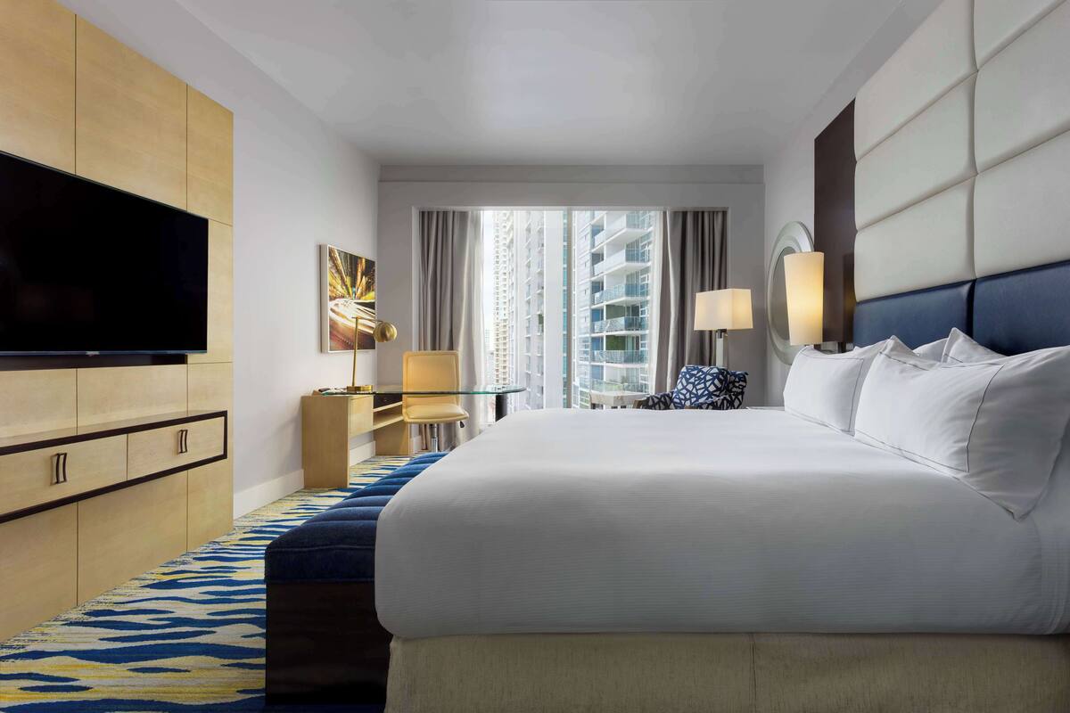 A hotel room with a large bed, a flat-screen TV, a desk with a lamp, and a view of high-rise buildings.