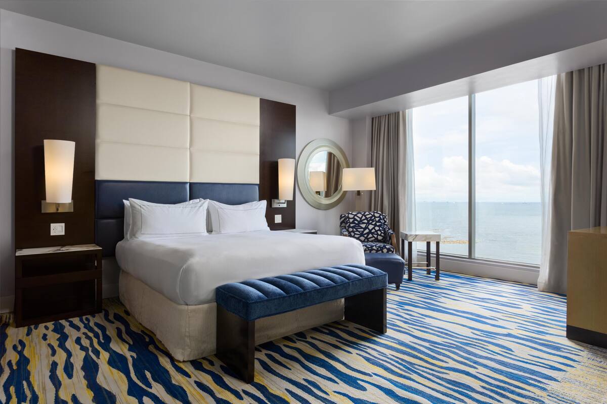 A hotel room with a large bed, a bench, a chair, a small table, a mirror, and a view of the ocean.