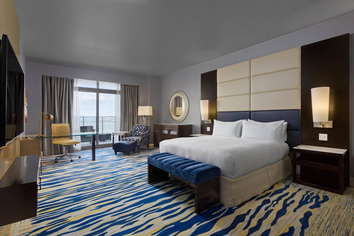A hotel room with a large bed, a desk with a chair, a round mirror, a sofa, and a view of the outdoors.