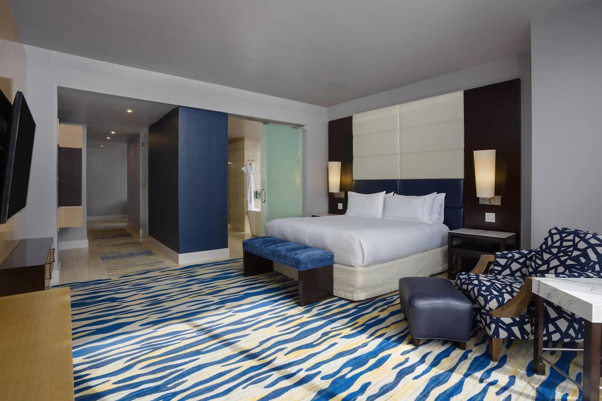A modern hotel room with a large bed, a desk, a chair, and a patterned rug.