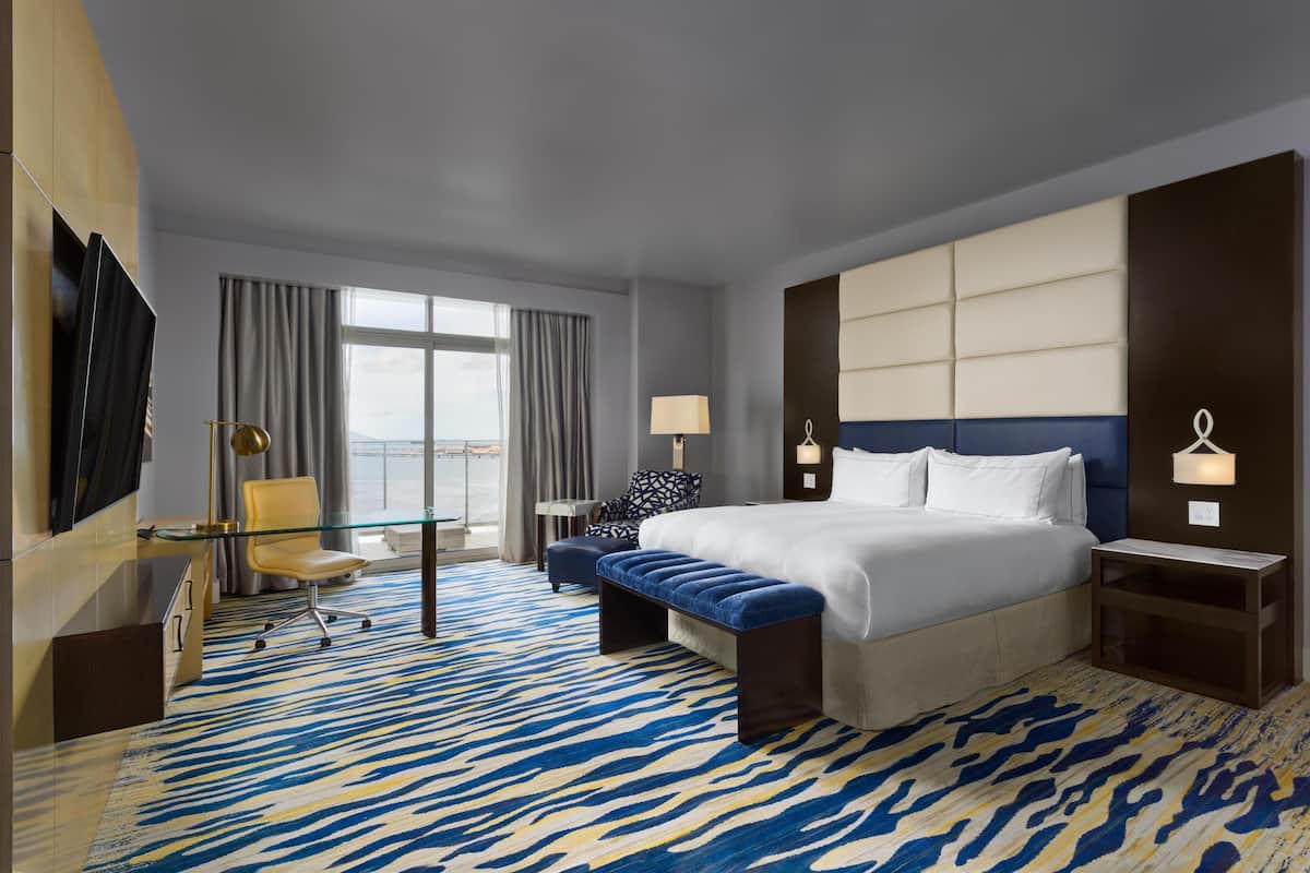 A hotel room with a large bed, a desk, a chair, a bench, and a TV.