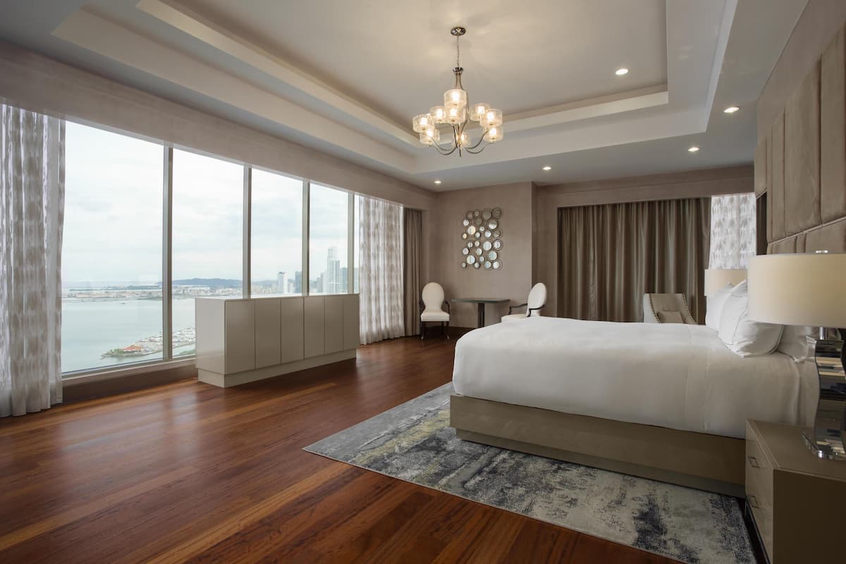 A modern hotel room with a large bed, a view of the city, a chandelier, and a small table with chairs.