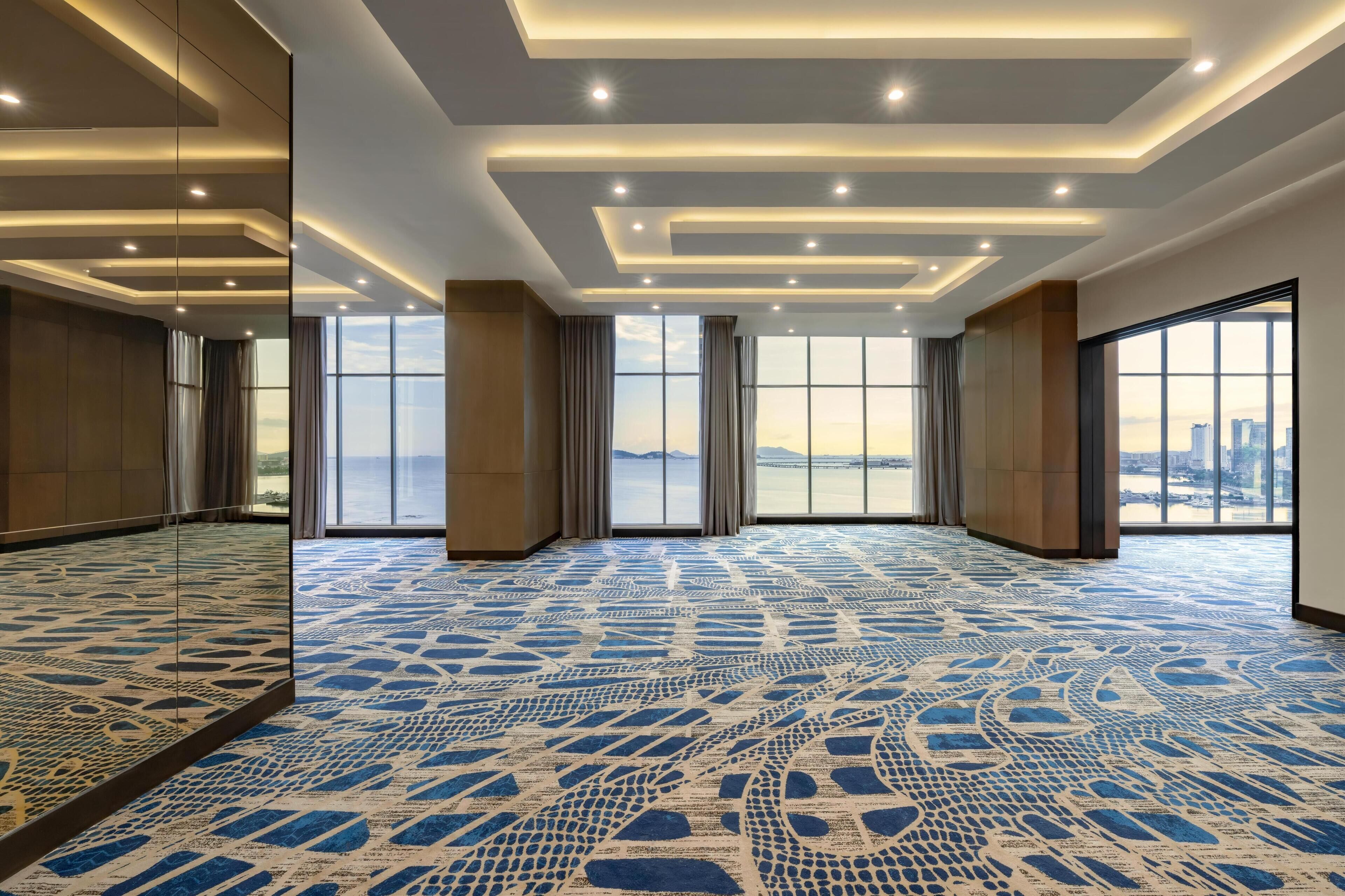 Une pièce spacieuse dotée d’un tapis à motifs, de grandes fenêtres avec des rideaux et d’un design de plafond moderne.