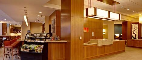 Un hall d’hôtel moderne avec un comptoir d’accueil central en bois, un espace salon et un présentoir de nourriture.