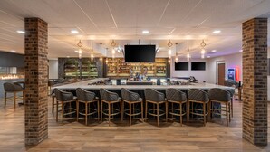 Uma área de bar com balcão comprido, vários bancos altos, uma tela de televisão e uma estante de bebidas bem abastecida.