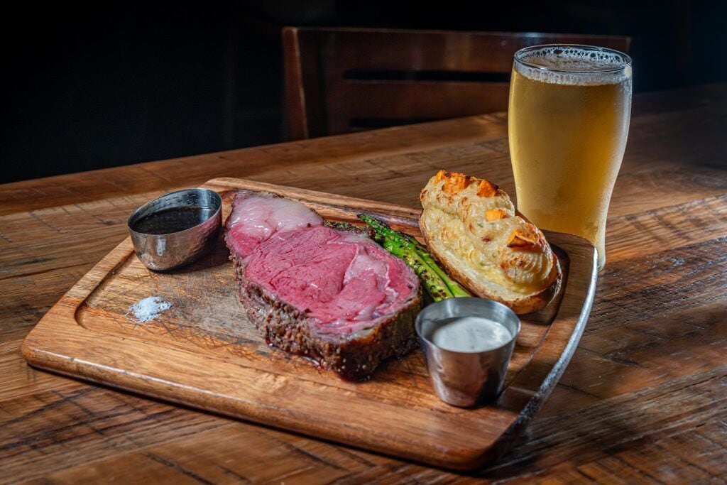 A wooden table with a glass of beer, a slice of meat, asparagus, and a small dish of sauce.