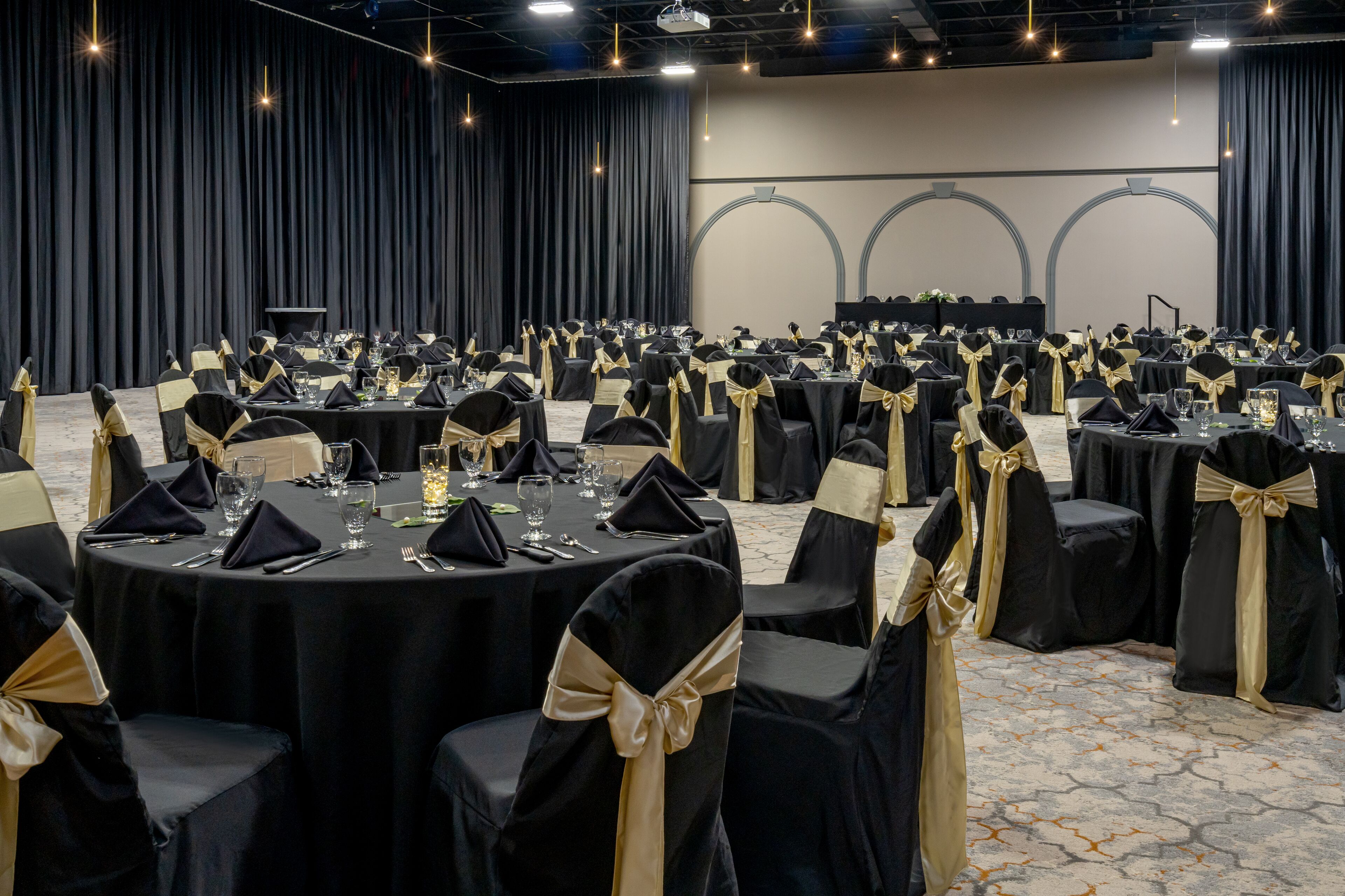 Un salón de banquetes con mesas redondas, manteles negros y sillas con cintas doradas.