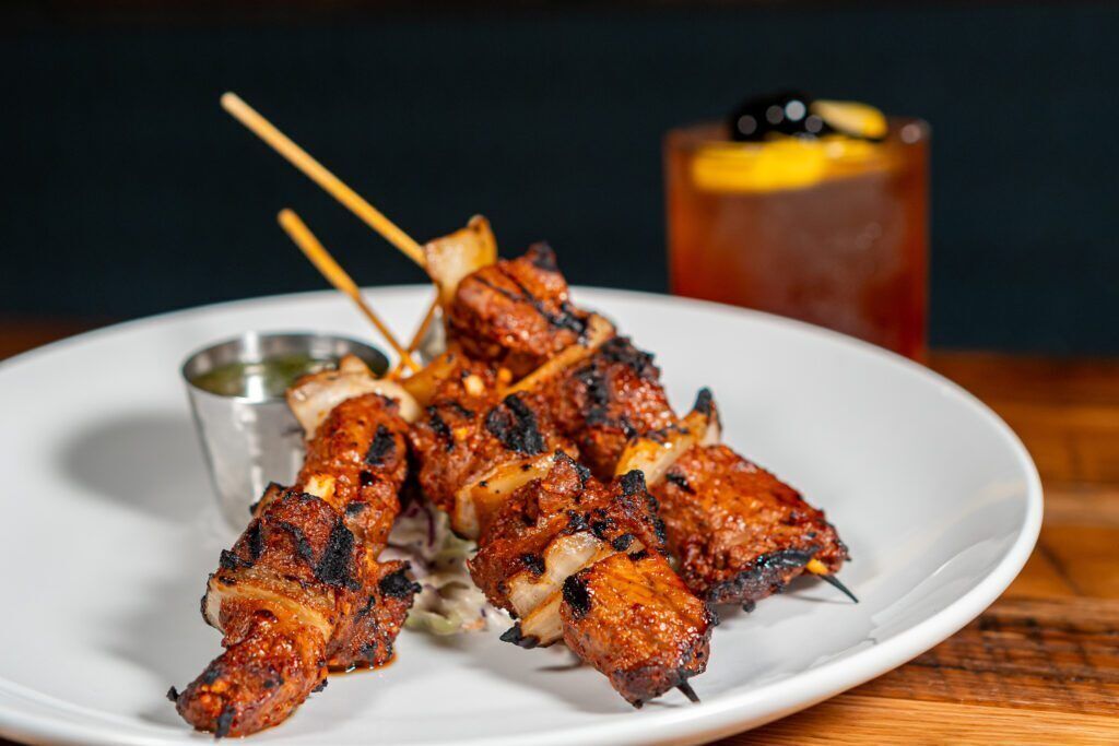 A plate of grilled chicken skewers with a side of sauce and a glass of iced tea.