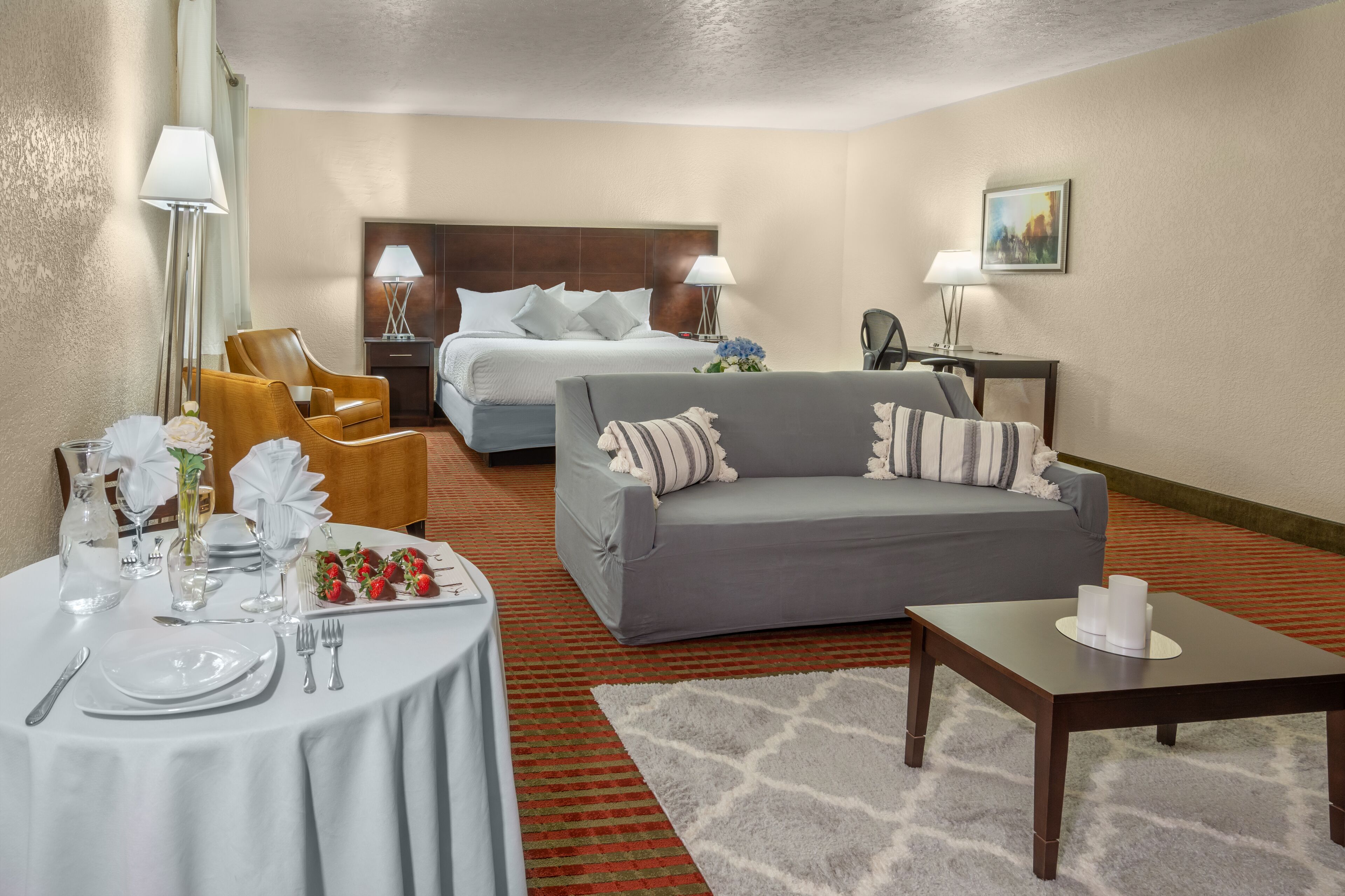 Habitación de hotel con cama, sofá gris, mesita con vela y zona de comedor con mesa dispuesta para dos.