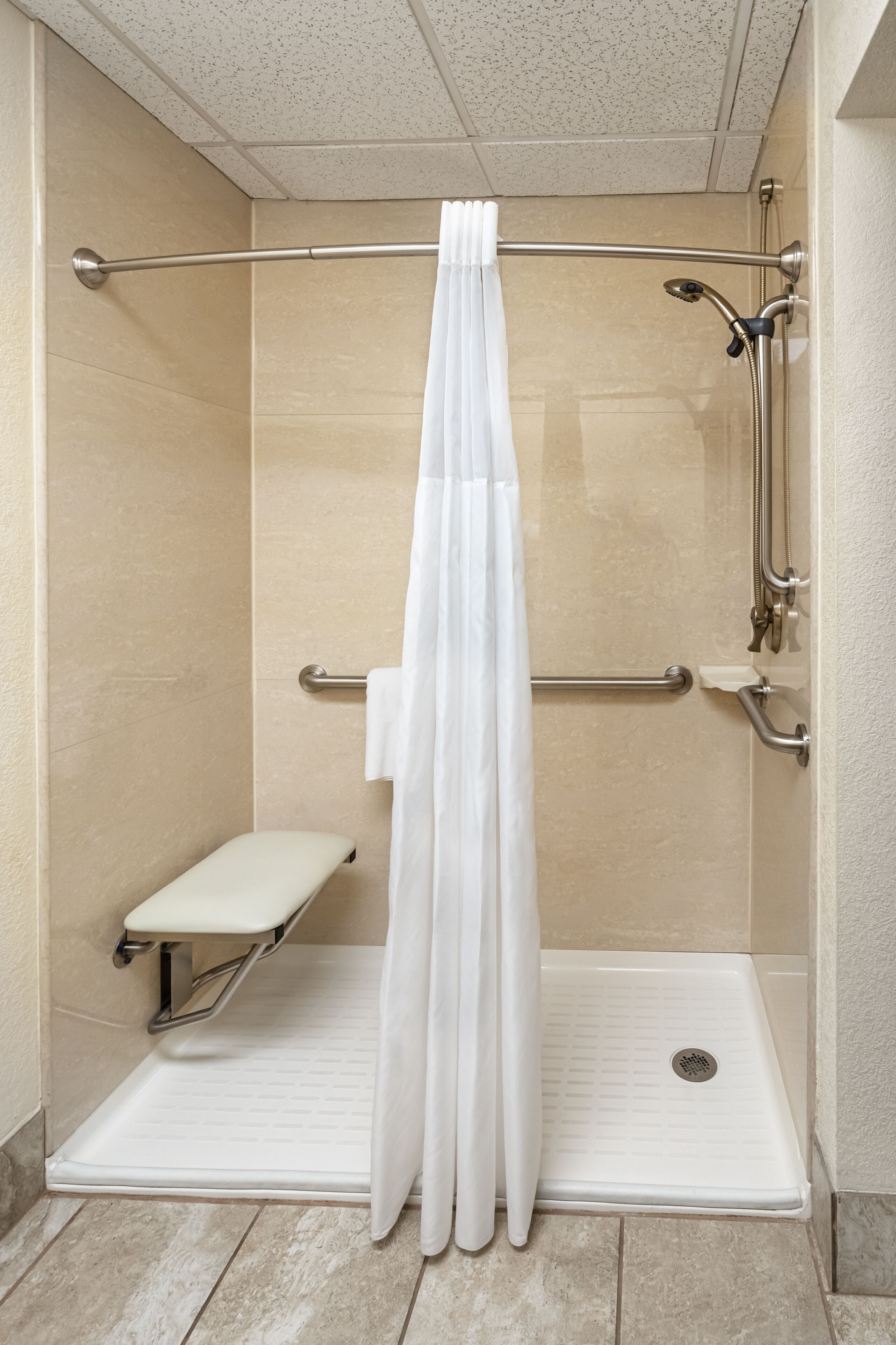 A shower area with a white shower curtain, grab bars, a bench, and a handheld showerhead.