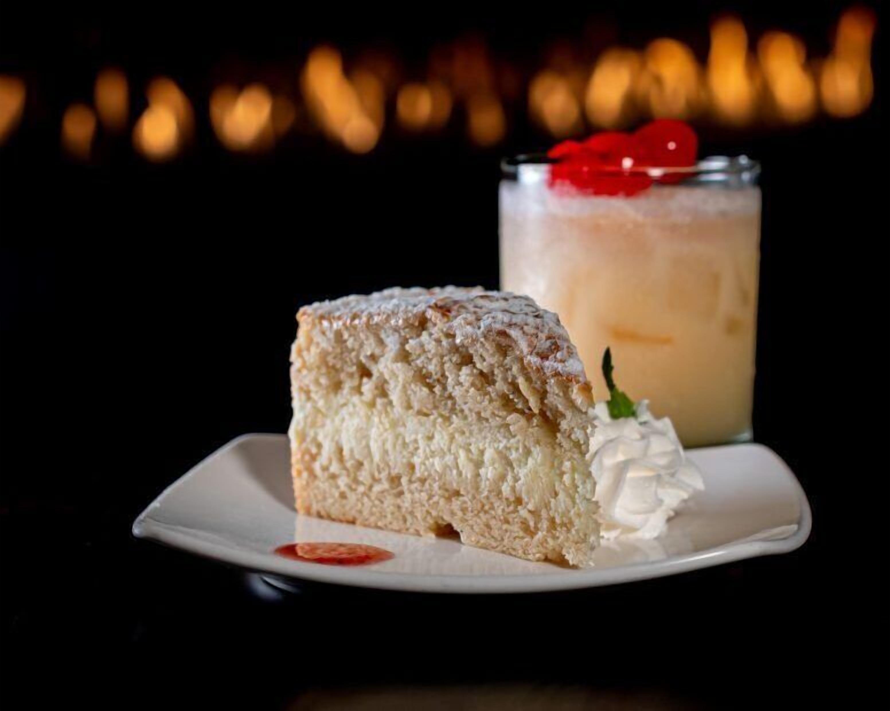 A slice of cake with whipped cream and a cherry on a white plate, with a glass of a creamy beverage in the background.
