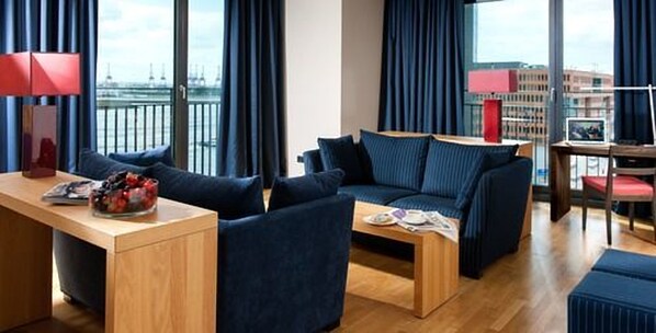 A modern hotel room with a dark blue sofa set, a wooden coffee table, and a view of the city through large windows. - Clipper Boardinghouse – Hamburg-Holzhafen (Hamburg)