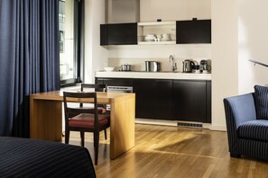 A modern hotel room with a wooden dining table, a single chair, a striped sofa, and a kitchen area with black cabinets and white countertops. - Clipper Boardinghouse – Hamburg-Holzhafen (Hamburg)