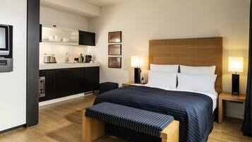Une chambre d’hôtel moderne dotée d’un grand lit, de tables de chevet avec des lampes, d’une tête de lit en bois, d’un banc, d’une kitchenette avec un téléviseur et d’un téléviseur à écran plat fixé au mur.