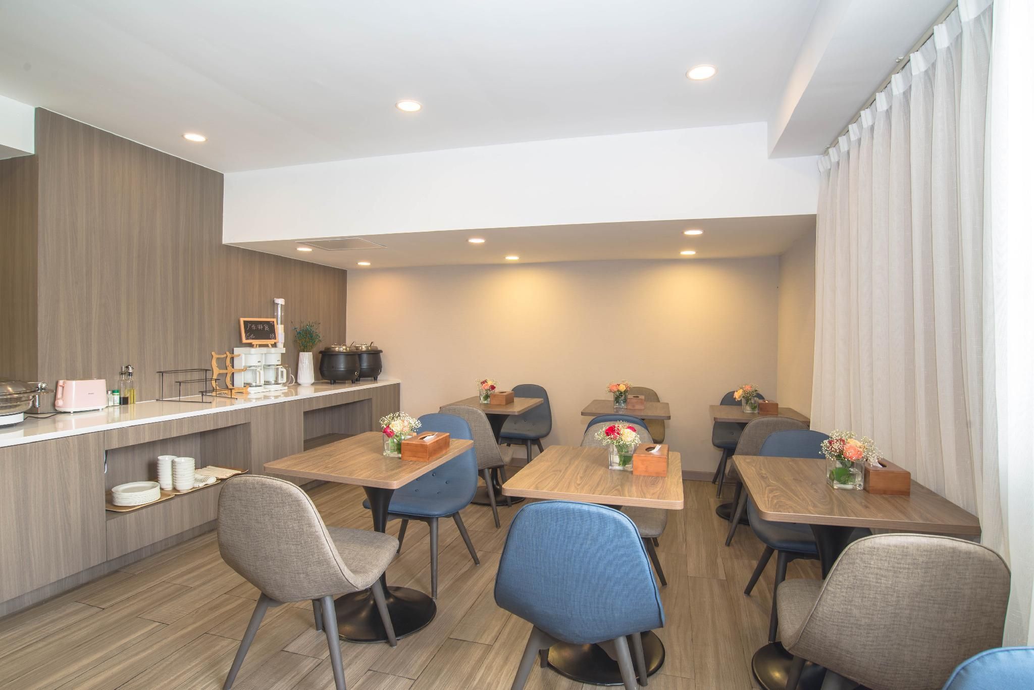 A modern dining area with wooden tables and chairs, a kitchen counter with appliances, and a large window with curtains.